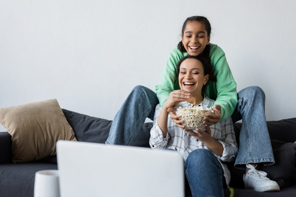 happy african american woman with teenage daughter watching movie on laptop near bowl of popcorn - Foto, afbeelding