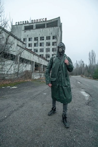 Мужчина в противогазе и костюм для защиты от радиации на фоне заброшенной гостиницы в Полесье в городе Припять. Припять, Украина - 28 ноября 2021 года. - Фото, изображение