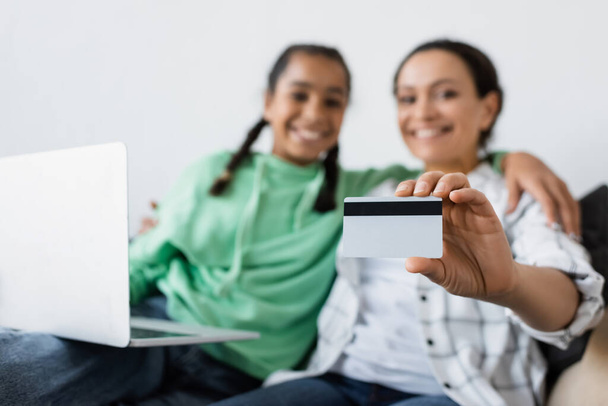 selective focus of credit card in hand of blurred african american woman sitting near laptop and teen daughter - Photo, Image
