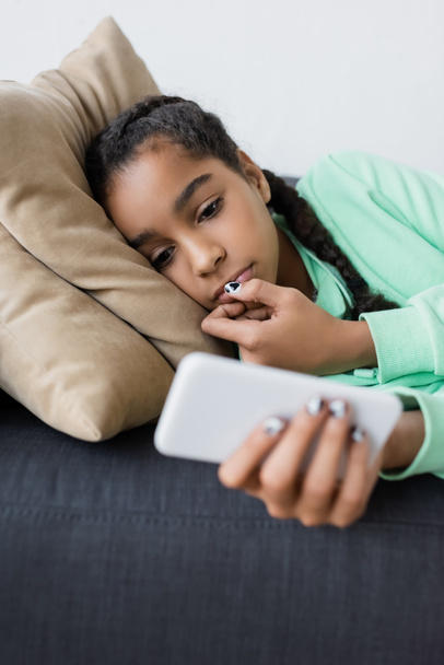 african american teenage girl looking at blurred smartphone while lying on couch at home - 写真・画像