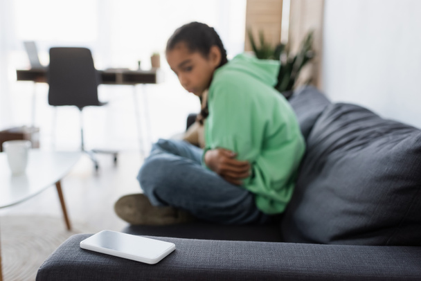 menina americana africana borrada de mau humor sentado no sofá e olhando para o smartphone com tela em branco - Foto, Imagem