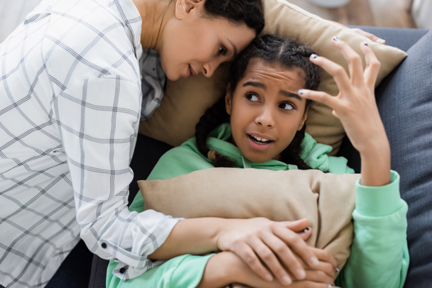 african american woman calming irritated daughter gesturing while lying on couch with pillows - Photo, Image