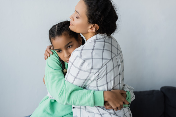 Afrikanisch-amerikanische Mutter mit geschlossenen Augen umarmt depressive Teenager-Tochter zu Hause - Foto, Bild
