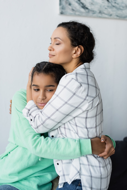 african american woman with closed eyes embracing frustrated daughter at home - Foto, imagen