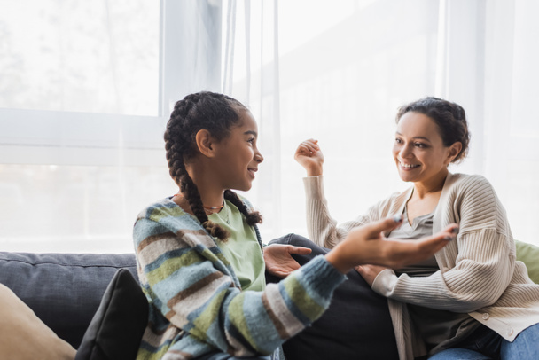 teenage african american girl pointing with hand while talking to smiling mother on sofa - Foto, Imagen