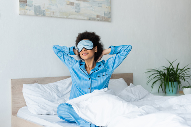 Χαρούμενη Αφροαμερικανή γυναίκα με πιτζάμες και μάσκα ύπνου να κάθεται στο κρεβάτι - Φωτογραφία, εικόνα