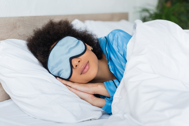 curly african american woman in blue pajamas and silk eye mask sleeping in bed - Foto, imagen