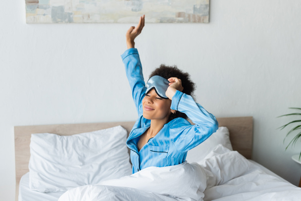 sleepy african american woman in pajamas adjusting sleeping mask and stretching while smiling in bedroom  - Fotó, kép