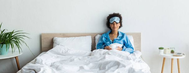 tevreden Afro-Amerikaanse vrouw in blauwe pyjama en slaapmasker met kopje koffie in bed, banner - Foto, afbeelding