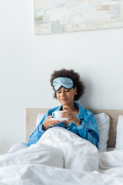 happy african american woman in pajamas and sleeping mask holding cup of coffee in bed - Foto, Imagen