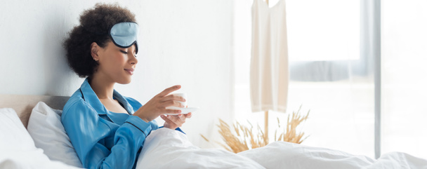 mujer afroamericana complacida en pijama y máscara para dormir sosteniendo taza de café en la cama, pancarta - Foto, Imagen