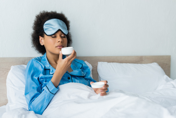mujer afroamericana en pijama y máscara para dormir sosteniendo contenedor y oliendo crema cosmética en la cama  - Foto, imagen