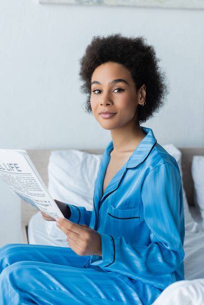 african american woman in pajamas holding travel life newspaper in bedroom  - Foto, imagen