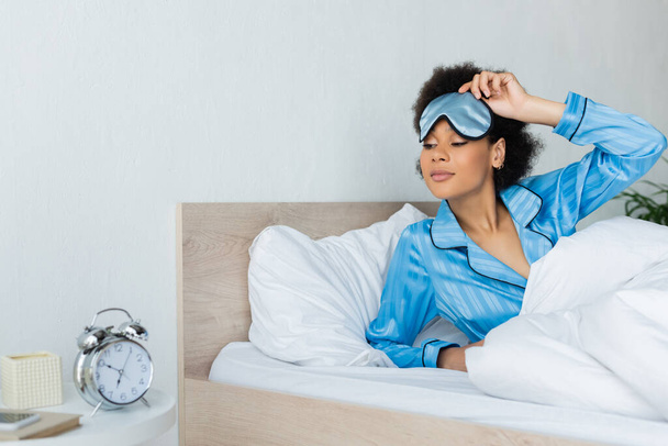 awake african american woman in pajamas adjusting sleeping mask while looking at alarm clock in bedroom - Foto, afbeelding