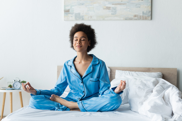 ευχαριστημένη Αφροαμερικανή με πιτζάμες που διαλογίζεται στο κρεβάτι. - Φωτογραφία, εικόνα