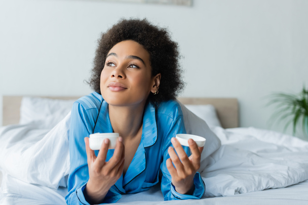 curly african american woman in pajamas lying on bed and holding container with face cream - Foto, Imagen