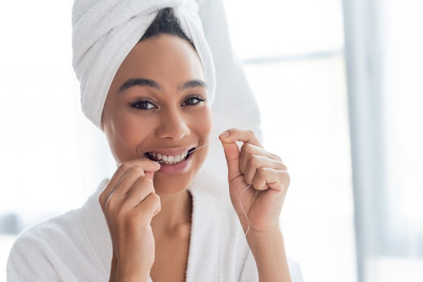 smiling young african american woman in bathrobe flossing teeth in bathroom - 写真・画像
