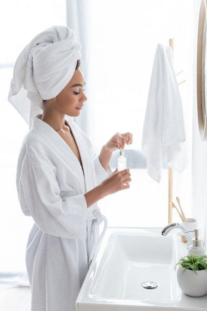 vista lateral de la joven afroamericana en una botella de algodón blanco con suero en el baño  - Foto, Imagen