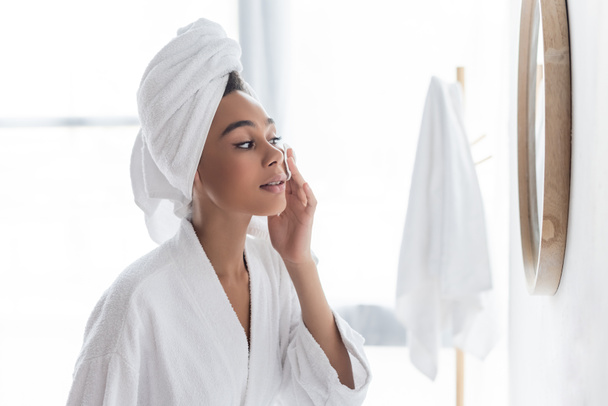 young african american woman in towel cleansing face with cotton pad near mirror - Fotografie, Obrázek