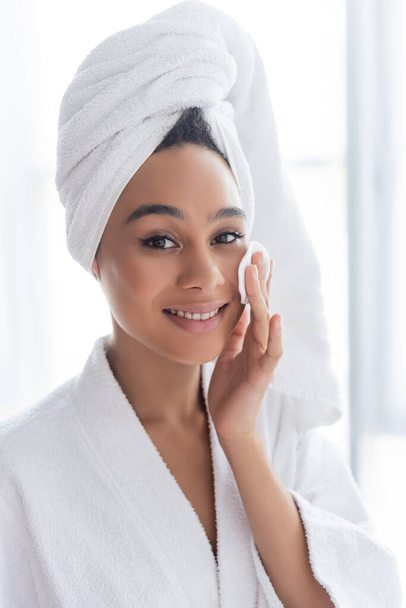 cheerful african american woman in towel cleansing face with cotton pad in bathroom  - 写真・画像
