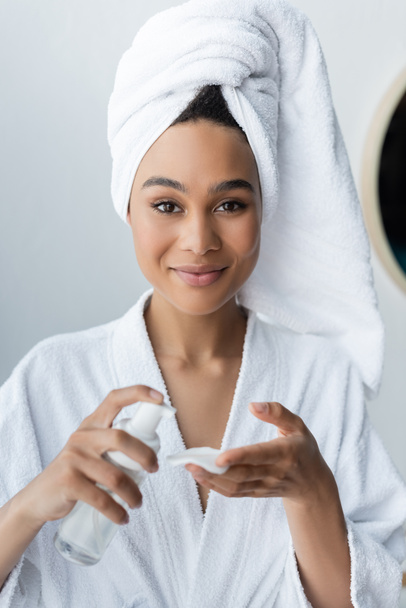 joyful african american woman in towel holding bottle with cleanser and cotton pad in bathroom  - 写真・画像