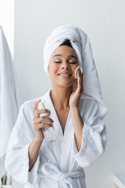 joyful african american woman in towel holding bottle and cleansing face with cotton pad - Foto, Bild