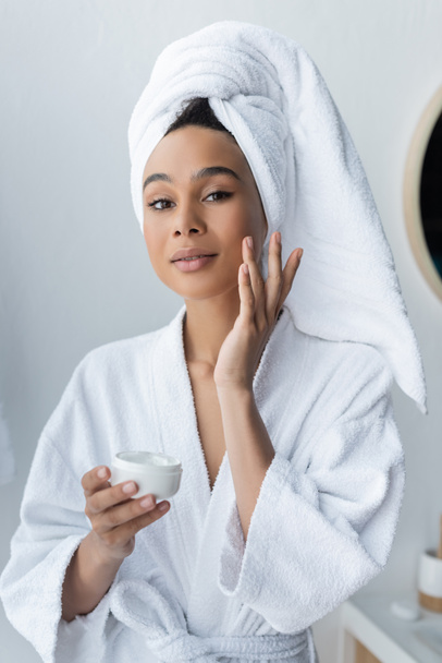 african american woman in bathrobe holding container and applying face cream in bathroom  - Фото, изображение