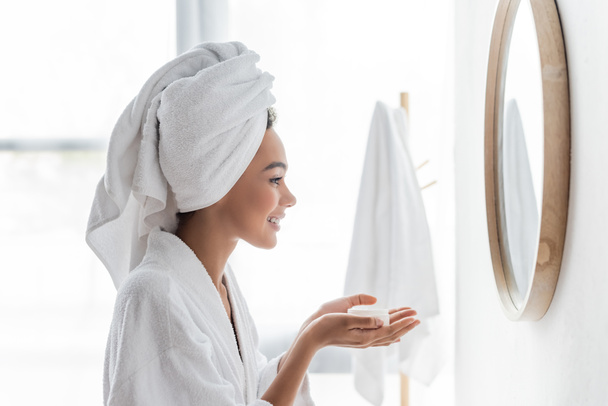side view of happy african american woman in bathrobe holding container with face cream and looking at mirror  - Photo, Image