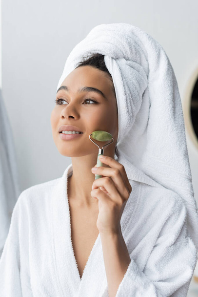 smiling young african american woman in bathrobe and towel massaging face with jade roller  - Фото, зображення