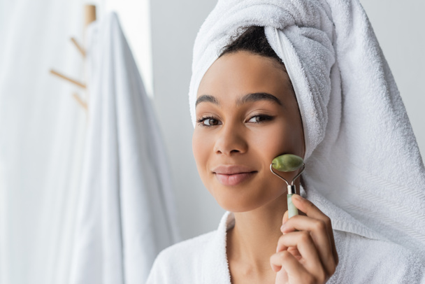 pleased african american woman in white towel massaging face with jade roller in bathroom  - Фото, изображение