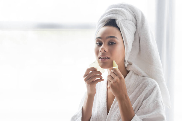 young african american woman in white towel massaging face with jade face scrapers - Photo, Image