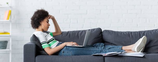 side view of curly african american woman sitting with laptop on couch, banner - Foto, immagini