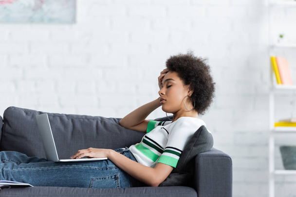 sleepy african american woman sitting with laptop on couch - Fotó, kép