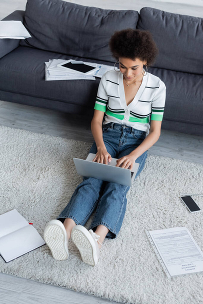 high angle view of african american freelancer using laptop and sitting on carpet  - Photo, Image
