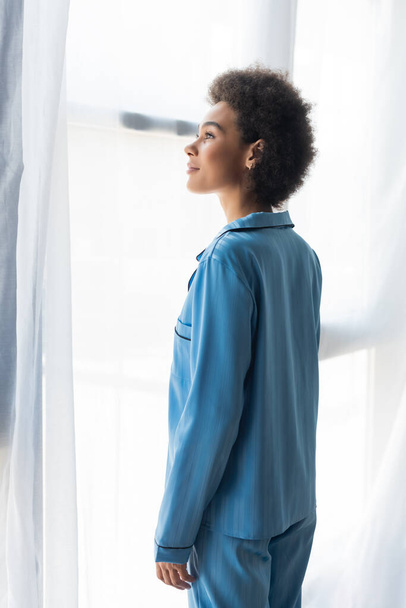 Boční pohled na africkou americkou ženu v modrém pyžamu stojící u záclon  - Fotografie, Obrázek