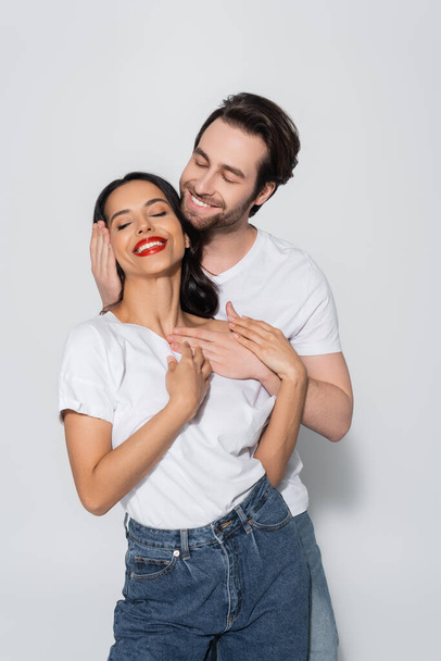 smiling man embracing happy brunette woman standing with closed eyes on grey - Foto, afbeelding