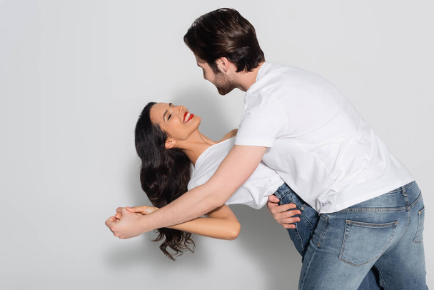 jovem homem de t-shirt branca dançando com mulher morena sorridente em cinza - Foto, Imagem