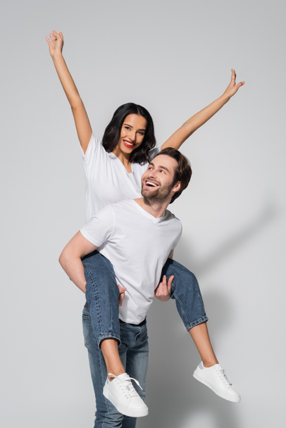 cheerful man piggybacking excited brunette woman in white t-shirt and jeans on grey - Zdjęcie, obraz
