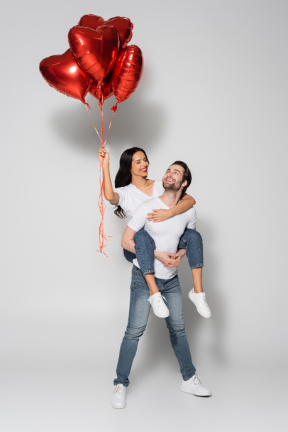 full length view of happy man piggybacking girlfriend with heart-shaped balloons on grey - Fotó, kép