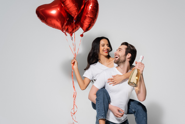 hombre sosteniendo botella de champán mientras que piggybacking mujer alegre con globos en forma de corazón en gris - Foto, imagen