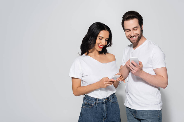 smiling brunette woman and bearded man in white t-shirts using smartphones on grey - 写真・画像