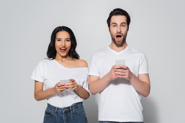 amazed couple in white t-shirts looking at camera while using smartphones on grey - Foto, Imagem