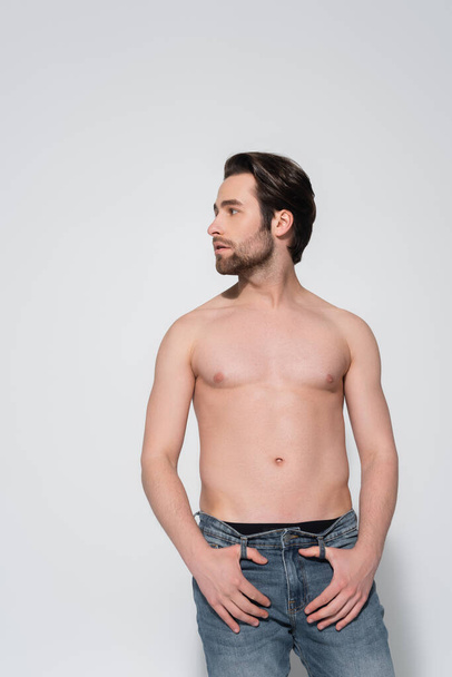 brunette shirtless man in jeans looking away while standing with thumbs in belt loops on grey - Fotó, kép