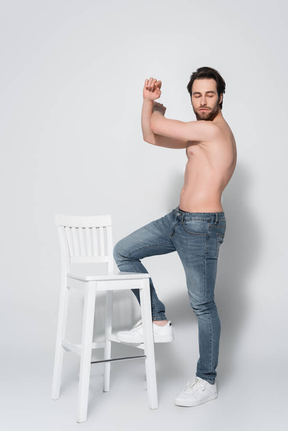 full length view of shirtless man in jeans posing near white chair on grey - Zdjęcie, obraz