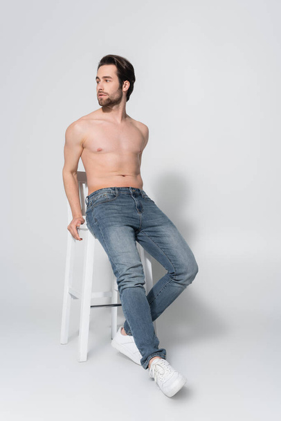 full length view of shirtless man in jeans looking away while posing on white chair on grey - Fotoğraf, Görsel