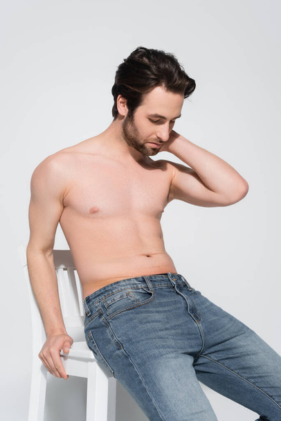 shirtless man in jeans touching neck while sitting on white chair on grey - Fotoğraf, Görsel