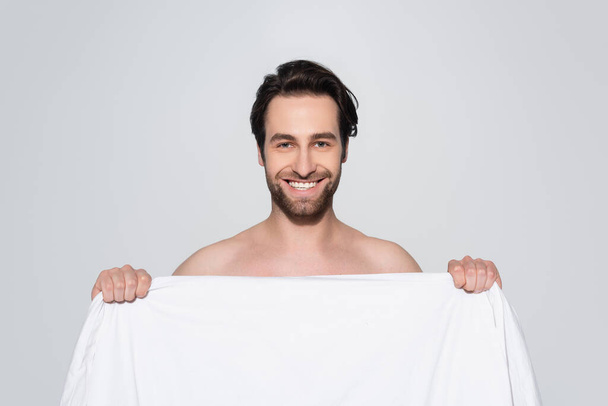 brunette shirtless man smiling at camera behind white blanket isolated on grey - Zdjęcie, obraz