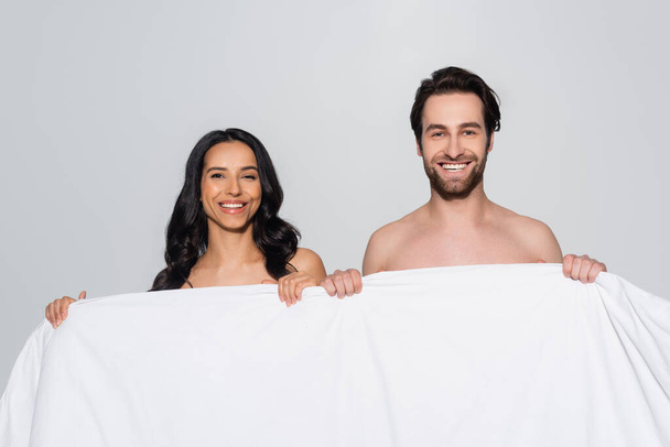 cheerful brunette couple smiling at camera while holding white blanket isolated on grey - Foto, Imagen