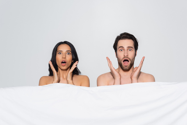 shocked couple with open mouths looking at camera near white blanket isolated on grey - Photo, Image
