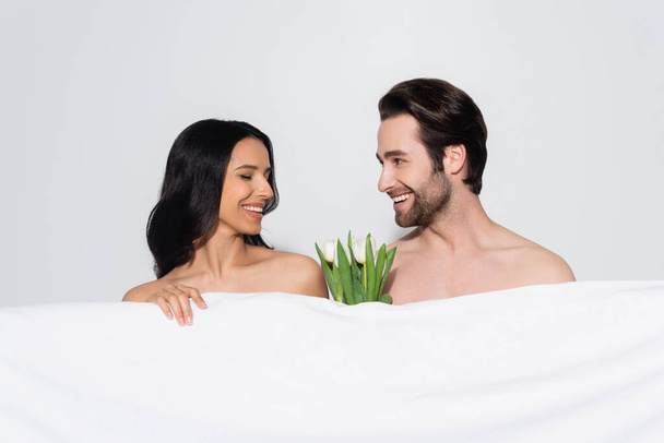 pleased lovers looking at each other near white blanket and tulips isolated on grey - Фото, изображение